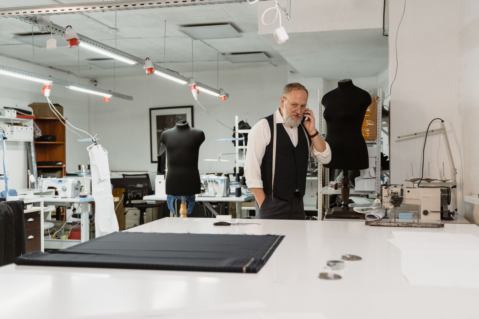 Un tailleur relance un client au téléphone depuis son atelier, entouré de ses machines à coudre et de ses mannequins