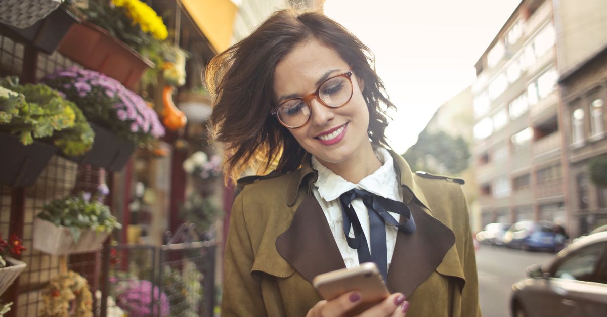 Une cliente souriante consulte son smartphone dans la rue, devant la devanture fleurie d'un commerce