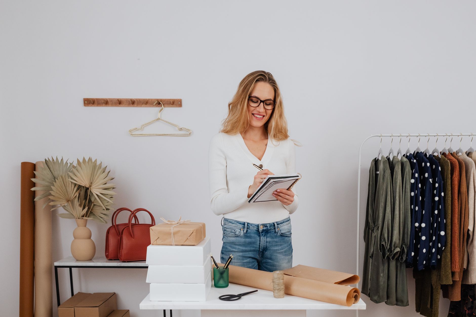 Une commerçante souriante planifie ses préparatifs dans sa boutique, entourée de paquets, de papier cadeau et d'un portant de vêtements