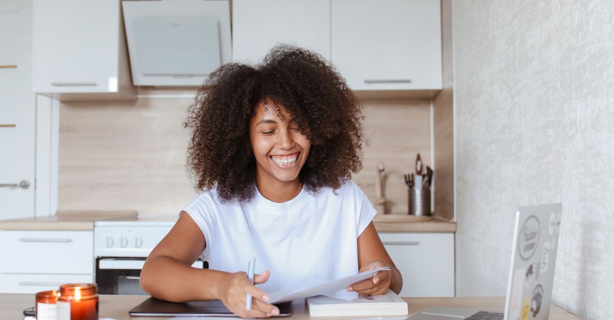 Une entrepreneure souriante prépare ses documents chez elle, ordinateur et carnet à portée de main